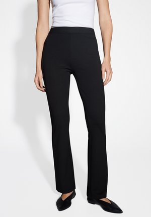 Frau trägt schwarze eng anliegende Bootcut-Hose mit elastischem Bund, kombiniert mit weißem ärmellosem Top und schwarzen spitzen Schuhen.