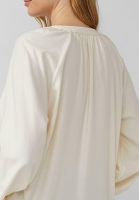 Vrouw met haar rug naar ons toe, gekleed in een losse, crèmekleurige top met lange mouwen en subtiele plooien bij de achterkant van de halslijn.