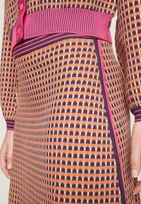 Abito in maglia con motivo a quadretti nei colori giallo senape, viola e rosa. Presenta una vita a coste rosa e bottoni decorativi.