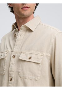 Beige overhemd met knopen, voorzien van twee borstzakken, gemaakt van gestructureerde stof, met metalen knopen en een klassiek kraagontwerp.