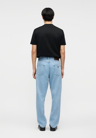 Άτομο που στέκεται με την πλάτη προς τα έξω, φοράει απλό μαύρο κοντομάνικο μπλουζάκι, ανοιχτόχρωμα μπλε τζιν με πίσω τσέπες και μαύρα παπούτσια.