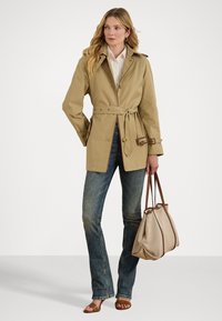 Manteau beige avec ceinture, comprenant une fermeture à boutons, des épaulettes et des accents marron ; porté sur un jean bleu évasé et des sandales marron.