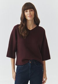 Bordeaux gebreide trui met een V-hals en korte mouwen, heeft een relaxed fit en een zachte textuur, gecombineerd met donkere spijkerbroek.