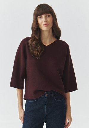 Maglione lavorato a maglia borgogna con scollatura a V e maniche corte, presenta una vestibilità rilassata e una texture morbida, abbinato a jeans in denim scuro.
