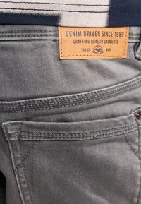 Grå denimshorts med et lædermærke med teksten "DENIM DRIVEN SINCE 1989." Har syningsdetaljer og åbninger til forlommer.