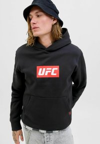 Giovane uomo che indossa un hoodie nero con il logo rosso della UFC, un cappello a secchiello nero e jeans grigio chiaro, che guarda di lato con le mani dietro la schiena.