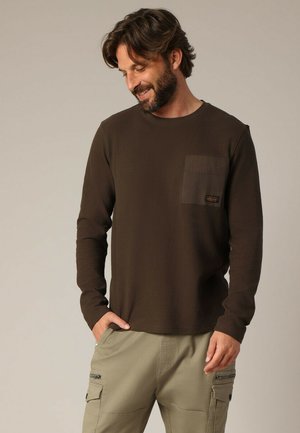 Bruine lange mouwen shirt gemaakt van zachte stof, met een borstzak in een donkere schaduw en een klein merklabel.
