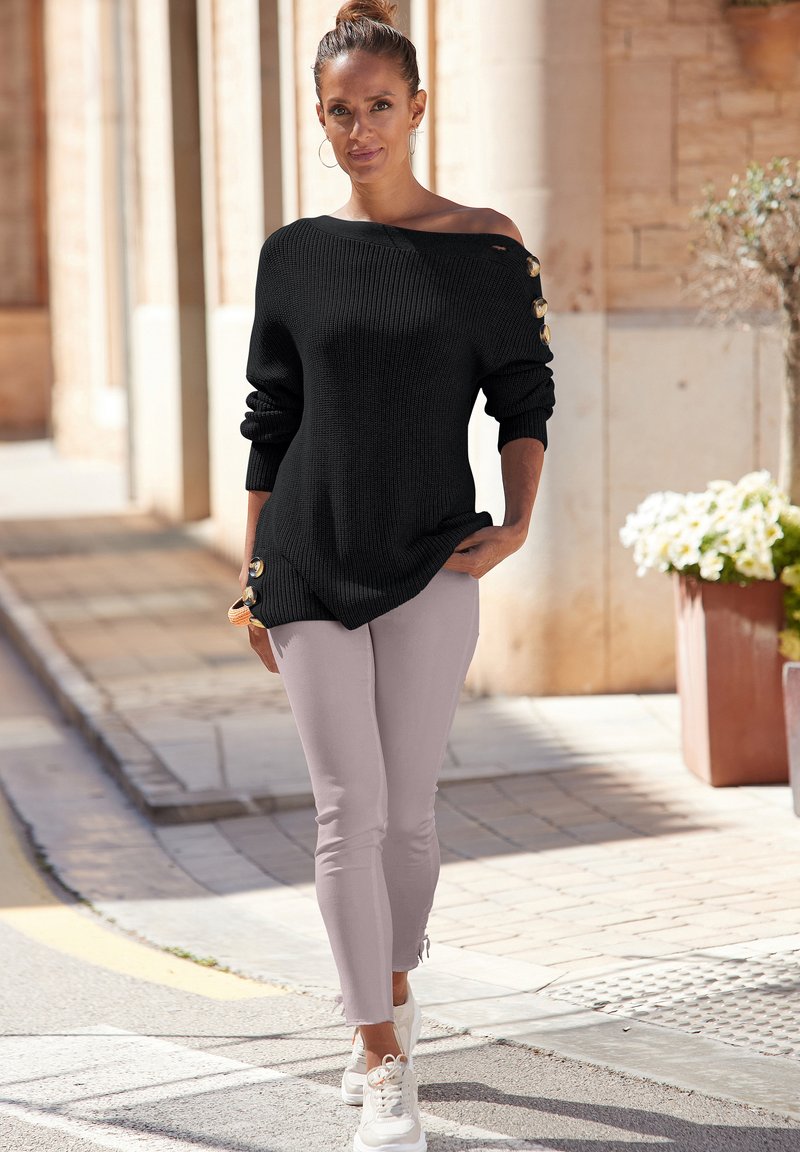 Zwarte gebreide trui met off-shoulder ontwerp en gouden knoopaccenten. Gecombineerd met lichtroze stretchbroek en witte sportschoenen.