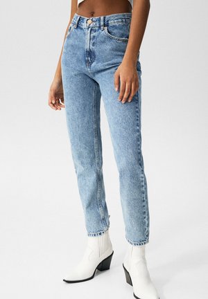 Femme portant un jean taille haute bleu clair et des bottines blanches à talons sur un fond uni.