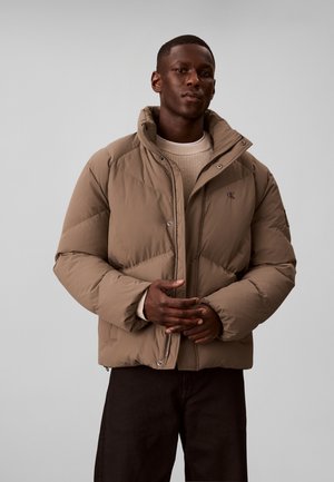 Homme portant une doudoune marron clair par-dessus un pull beige et un pantalon noir, debout les mains jointes devant un fond gris uni.