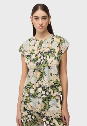 Donna con capelli lunghi e scuri, che indossa orecchini a cerchio dorati, una maglia a maniche corte con motivo floreale e pantaloni coordinati con motivo floreale, davanti a uno sfondo chiaro semplice.