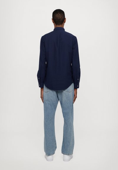 Donkerblauw overhemd met lange mouwen en een ronde zoom, knoopmanchetten en een rugpas, gecombineerd met lichtblauwe, losse jeans en witte sneakers.