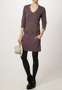 Vestito in maglia viola con scollatura a V e maniche a tre quarti. Presenta tasche con zip nere ed è abbinato a collant neri e stivali con tacco.