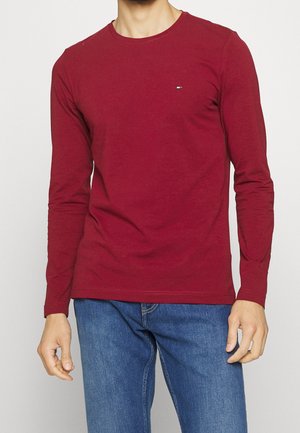 Homme portant une chemise rouge à manches longues unie avec un petit logo sur la poitrine et un jean bleu, debout devant un fond uni.