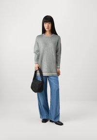 Maglione grigio oversize con strisce grigie chiare ai polsini, abbinato a jeans larghi blu chiaro. Tiene in mano una borsa a tracolla di tessuto nero intrecciato.