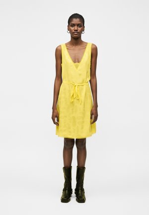 Γυναίκα με κίτρινο αμάνικο φόρεμα σε σχήμα V με κορδόνι στη μέση, συνδυασμένο με μαύρες μπότες μέχρι τη μέση της γάμπας, στέκεται μπροστά σε απλό φόντο.