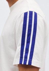 Λευκό κοντομάνικο πουκάμισο με τρεις παράλληλες μπλε ρίγες που τρέχουν κάθετα στην εξωτερική πλευρά του μανικιού.