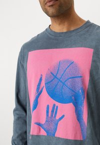 Langermet grå topp med grafisk design i rosa og blå farger. Designet viser basketballhender som strekker seg etter en ball.