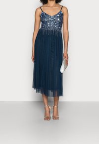 Vestido azul marino adornado con un corpiño ajustado de cuentas, falda de tul fluida y tirantes de espagueti ajustables. Se complementa con accesorios plateados.