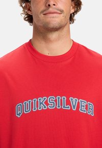 Camiseta roja de manga corta con escote redondeado que presenta "QUIKSILVER" en grandes letras azules en el pecho, fabricada en material de algodón.