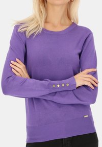 Fioletowy sweter z dzianiny, z okrągłym dekoltem, długimi rękawami i złotymi guzikami na mankietach. Gładka tekstura i dopasowany krój.