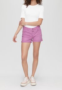 Mujer con una camiseta blanca de manga corta, pantalones cortos rosa con dobladillo, y zapatillas rosa claro y blancas, de pie con los brazos cruzados sobre un fondo blanco.