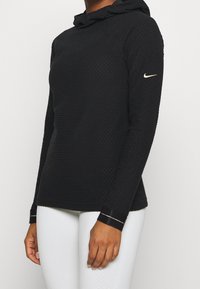 Maglia nera con cappuccio, maniche lunghe e motivo a nido d'ape, con accentuazione del logo sulla manica e polsini a contrasto con dettagli di cucitura.