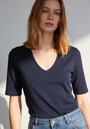 Frau mit schulterlangem hellbraunem Haar, die ein marineblaues V-Ausschnitt-Kurzarmshirt und blaue Jeans trägt, steht vor einem hellen Hintergrund.