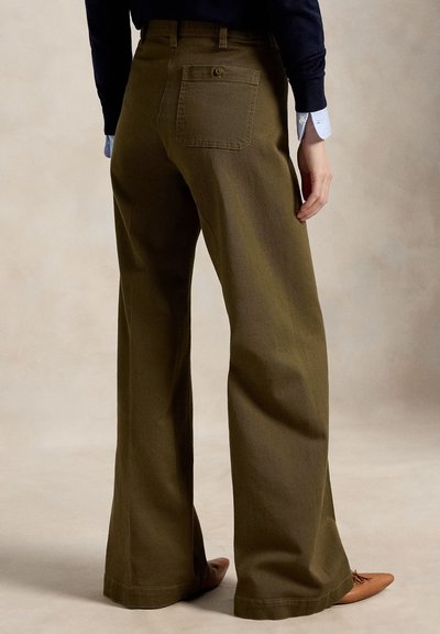 Person trägt weite olivgrüne Hosen und braune spitze Schuhe, steht vor einem neutralen Hintergrund.