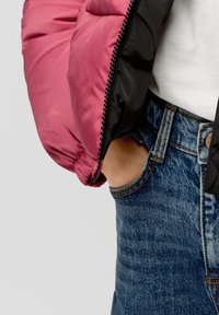 Veste matelassée avec un extérieur rose et des manches noires, munie d'une fermeture éclair, associée à un jean en denim bleu et à une chemise blanche.
