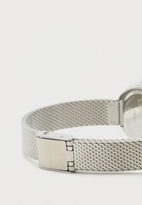 Ατσάλινο ρολόι με ασημένιο πλέγμα ιμάντα και πόρπη από ανοξείδωτο χάλυβα, με στρογγυλή κασέτα και γυαλιστερή επιφάνεια, με το όνομα της μάρκας.