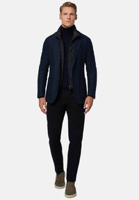 Giacca in misto lana blu navy indossata sopra un gilet nero con zip, abbinata a pantaloni neri e stivaletti color crema; design semplice con vestibilità su misura.