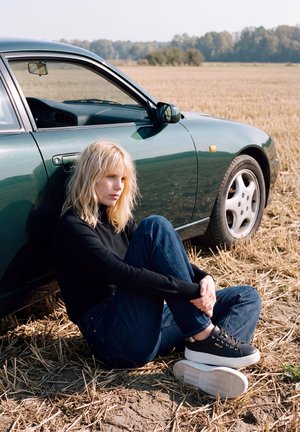 Une femme blonde est assise sur du chaume sec, à côté d’une voiture vert foncé, dans un champ moissonné.