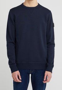 Mörkblå sweatshirt i mjukt tyg, med rundad hals, långa ärmar, ribbad manschett och en liten logopatch på vänster ärm.