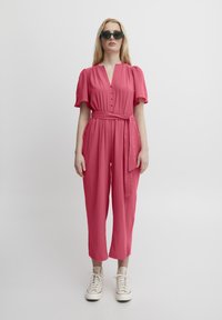 Gioconda rosa lampone con scollo a V, chiusura con bottoni, maniche corte e cintura da annodare. Indossata con sneakers bianche, caratterizzate da una texture liscia.