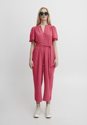Jumpsuit rose framboise avec un décolleté en V, fermeture à boutons, manches courtes et ceinture à nouer. Porté avec des baskets blanches, présentant une texture lisse.