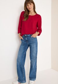 Rød bluse med lange ærmer og rund halsudskæring; blå, vide jeans med en falmet tekstur; hvide sneakers med røde accenter.