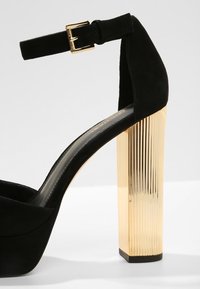 Chaussure à talon haut en daim noir avec un talon bloc épais en métal doré strié. Dotée d'une fine sangle à la cheville ajustable. Texture lisse.