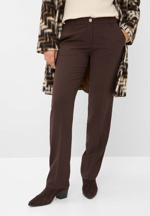 Henkilö, jolla on päällään tummanruskeat suorat housut, beige ribbineulepusero, kuvioitu ruskean ja valkoisen yhdistelmäinen takki sekä tummanruskeat nilkkurit korolla.