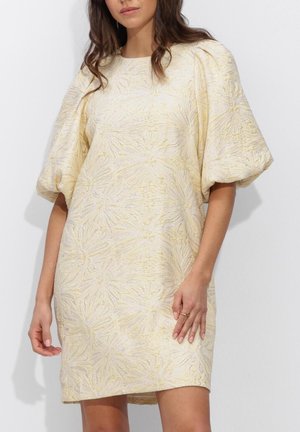 Vrouw die een lichtgeel getextureerde jurk draagt met korte pofmouwen en een bloemmotief, staand tegen een effen lichte achtergrond.