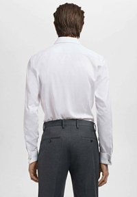 Camisa blanca de vestir con mangas largas, que presenta un cuello estándar. Combinada con pantalones oscuros de color gris con dos bolsillos traseros y detalles de botones.