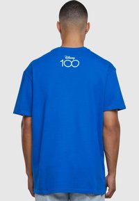 Blå bomull T-skjorte med rund hals, korte ermer og liten hvit Disney 100-logo øverst på ryggen. Myk tekstur og avslappet passform.