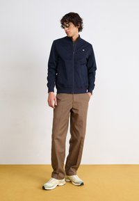 Marineblå zip-jakke med ståkrave, to sidelommer, kombinert med lysebrune bukser og kremfargede sneakers. Solbriller med grønne linser.