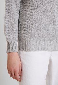 Pull gris tricoté avec un motif ondulé texturé, poignets et ourlet côtelés, associé à un pantalon blanc. Gros plan sur le bras et les détails du tissu.