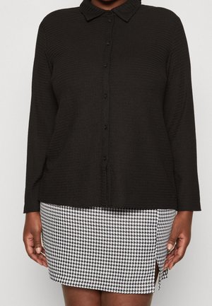 Person wearing a black textured button-up shirt and a black-and-white houndstooth skirt with a small front slit.
