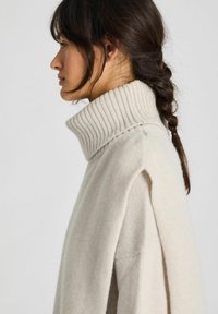 Mujer con cabello oscuro trenzado, vistiendo un suéter de punto color crema con cuello alto, mirando hacia la izquierda sobre un fondo claro y sencillo.