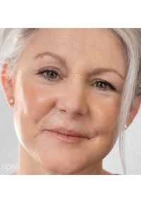Gros plan sur le visage d'une femme mature avec une peau claire, des cheveux argentés, des rides subtiles et des lèvres douces. Elle porte des boucles d'oreilles en perles et un maquillage neutre.