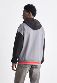Sweat à capuche avec des panneaux gris et noir, une bande rouge en accent au bas, texture douce et coupe décontractée. Porté avec un jean gris clair.