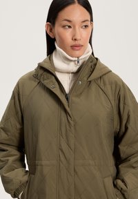 Oliivivihreä tikattu takki hupulla, jossa on vetoketjusulku ja napit. Päälle puettuna korkeakauluksinen kerma-valkoinen neuletakki.