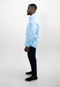 Lichtblauw overhemd met lange mouwen, gecombineerd met donkere broek en zwarte instapschoenen, gezien in profiel tegen een witte achtergrond.